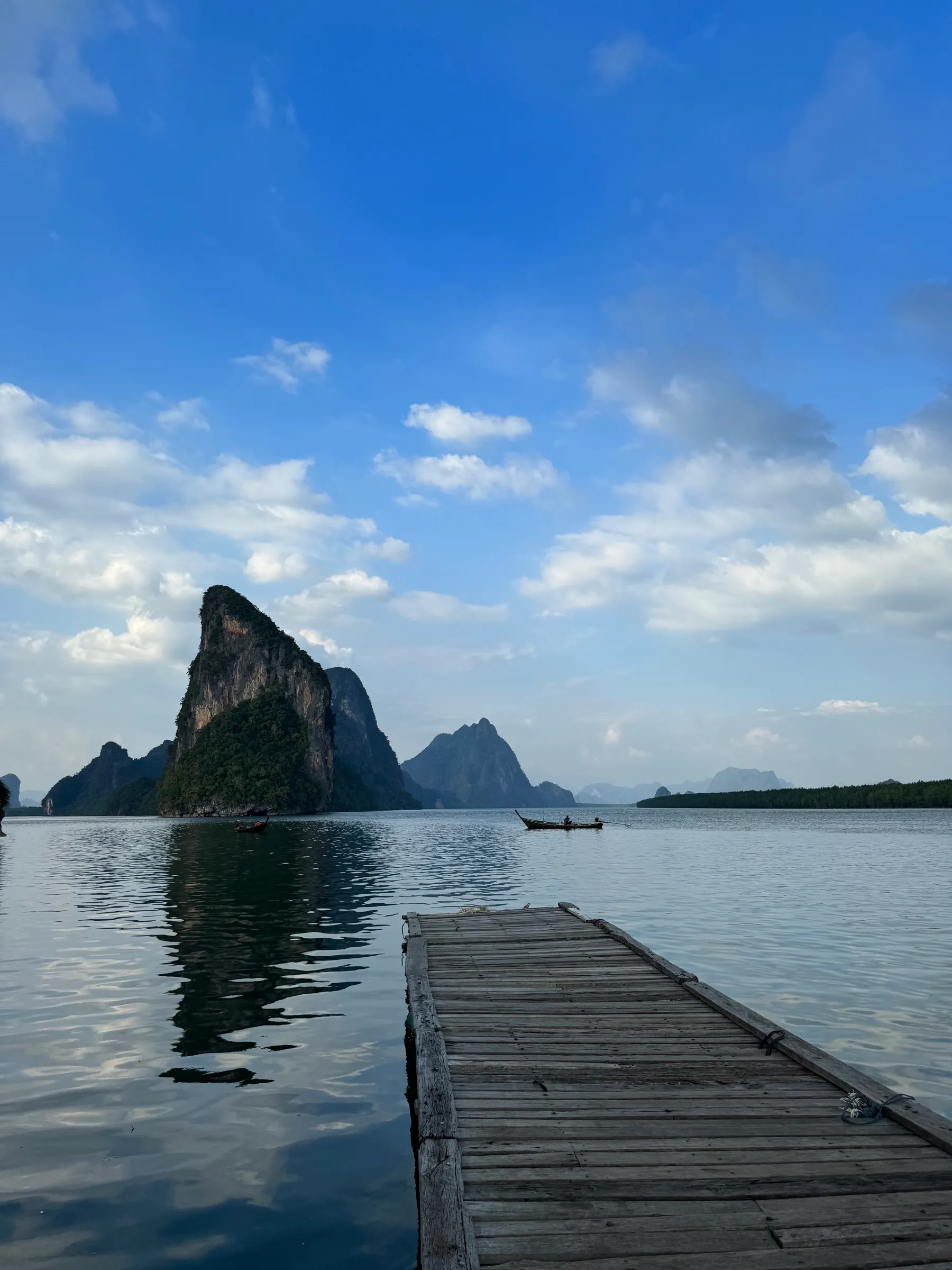 A wooden dock stretches toward the karst peaks of a calm bay, beneath a vast blue sky